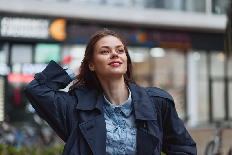 Smiling young woman wearing a denim shirt and dark jacket stands outdoors on an urban street, looking up with a joyful expression against a blurred city backdrop. Soft lighting adds warmth.