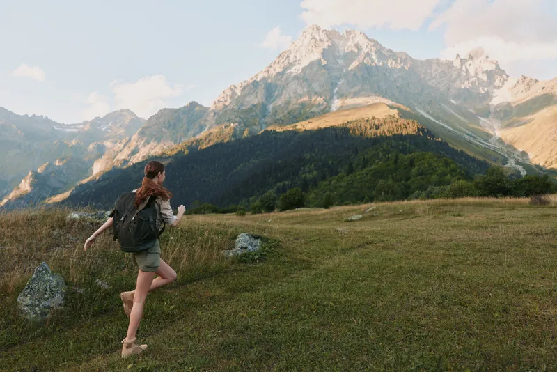 Woman hiking outdoors in nature with backpack walking on grassy field near mountains under blue sky, active lifestyle, adventure, freedom, summer day