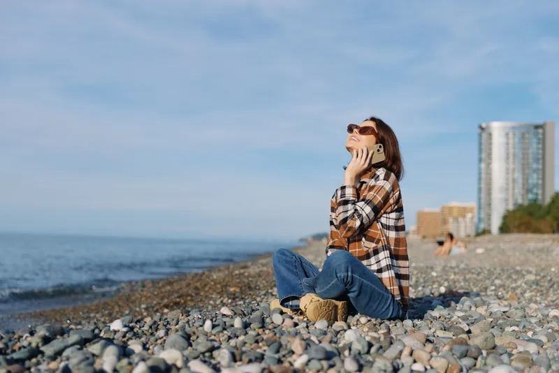 Woman on a pebble beach talks on her phone, casual plaid shirt and jeans, sea breeze, city skyline in the background, bright sunny day, relaxed outdoor moment, with waves and horizon