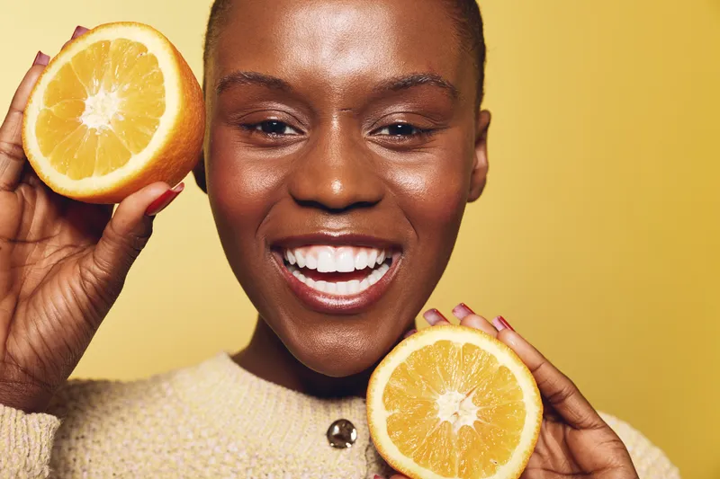 Smiling dark skinned African woman holding two orange halves near face, wearing beige sweater. Concept for healthy lifestyle, vitamin C, skincare, wellness and natural beauty on yellow background.