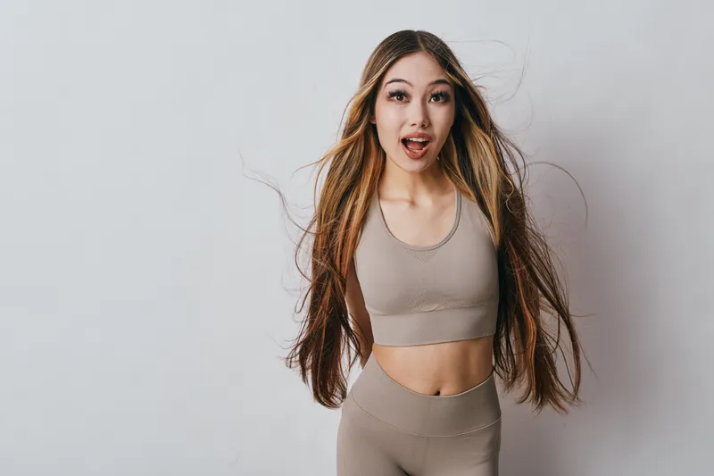 Woman with long hair in beige sportswear standing against white background. Female model with expressive surprised face, fitness outfit and casual posture for lifestyle and health.
