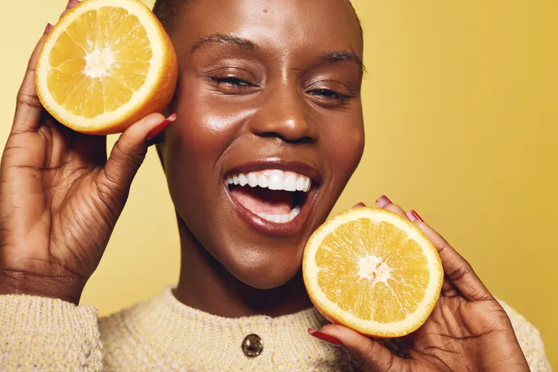Happy African woman with dark skin smiling broadly while holding two halves of an orange close to her face on yellow background. Concept of healthy lifestyle, vitamin C, and natural beauty.