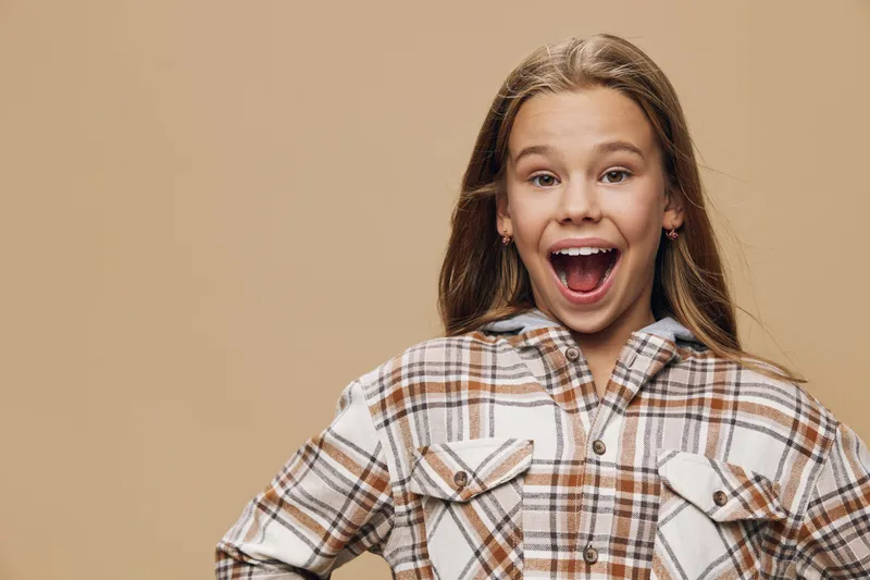 Girl with surprised expression and open mouth wearing checkered shirt on beige background. Child portrait with long hair and casual outfit in studio environment.
