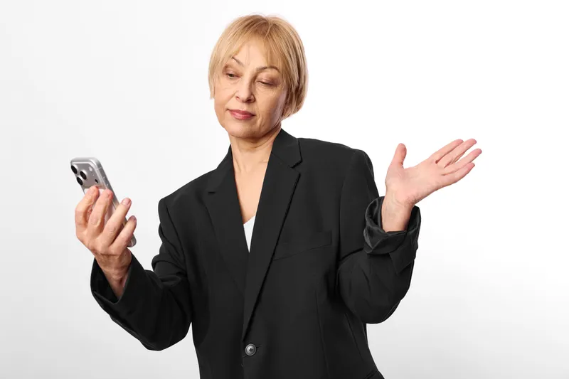 Woman in business suit holding smartphone, confused gesture with one hand raised, speaking without sound, isolated studio background, neutral expression, copy space, for ads today