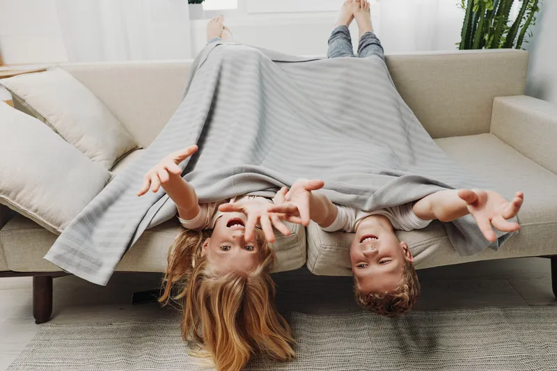 Two happy children lying upside down on a sofa covered with a striped blanket, playing and reaching out with their hands in a bright living room. Concept of childhood, fun, and family time.