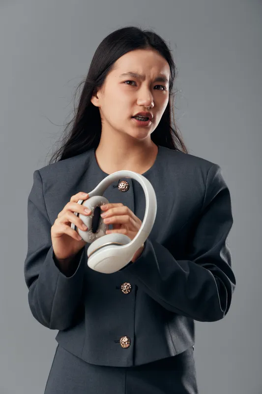 Woman holding headphones with a frustrated expression in gray suit against gray background. Young female showing emotion, technology accessory and casual style with long black hair.