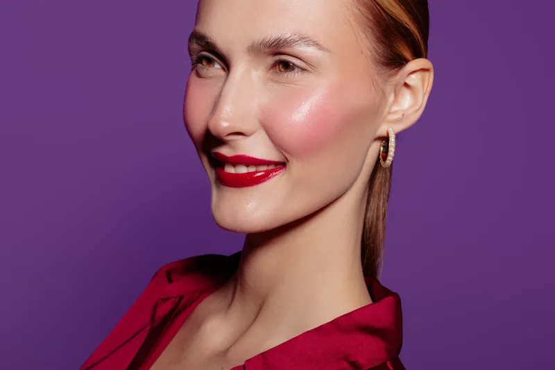 Smiling woman with red lipstick and shiny skin poses on purple background. Close-up portrait of confident female model with earrings and red blouse. Concept of beauty and makeup.