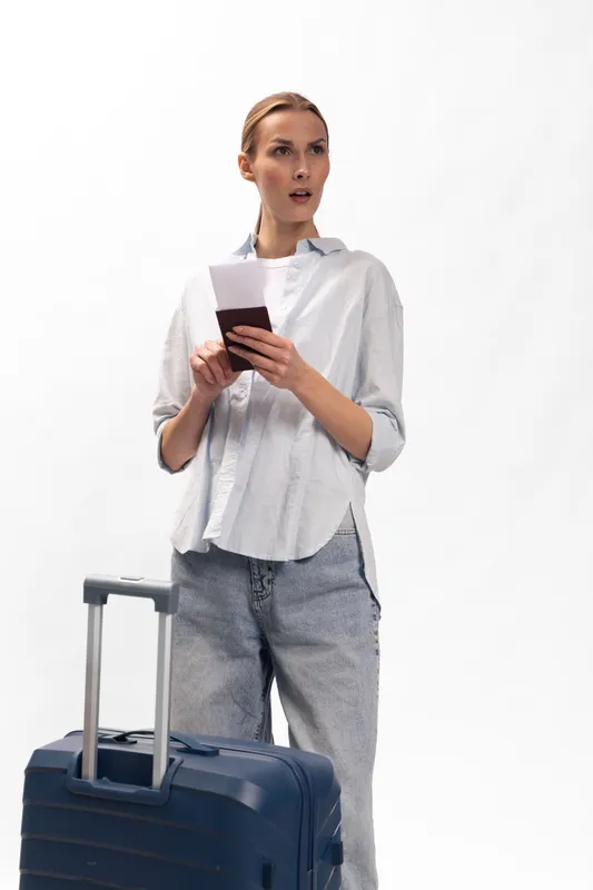 Young woman traveler with suitcase holds passport and phone looks surprised. Casual outfit on white background. Concept for travel, trip, tourism, and unexpected situation.
