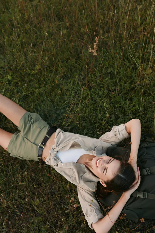 Young woman lying on grass with eyes closed and hands behind head, enjoying nature and relaxation outdoors. Casual outfit and peaceful mood in natural light.