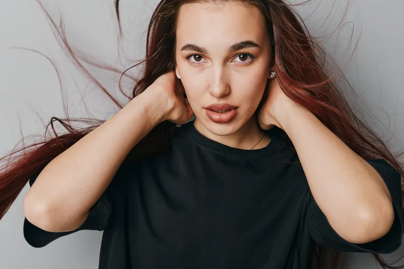 Young woman with long hair wearing black shirt posing with hands behind neck looking at camera against neutral background. Female model portrait with natural makeup and casual style.
