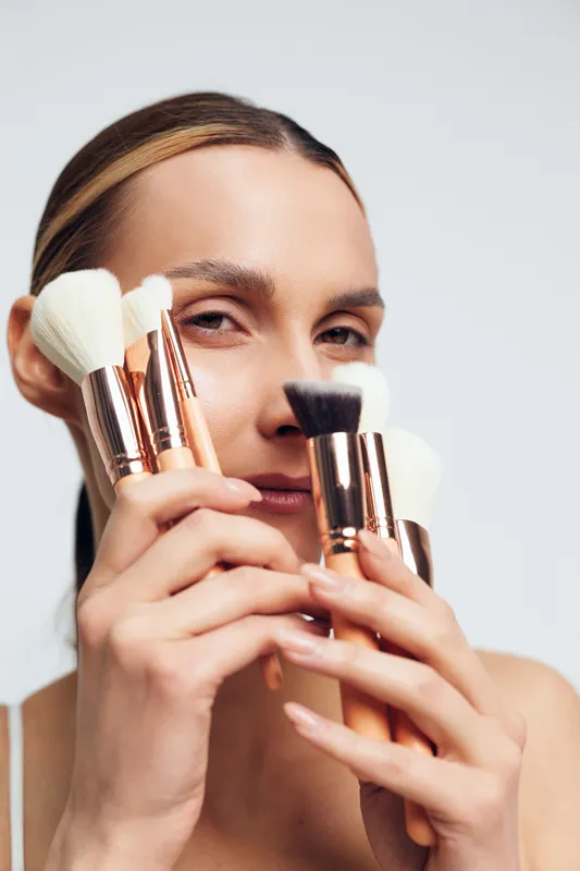 A woman holds several makeup brushes close to her face with a neutral expression. The image highlights beauty tools and cosmetics in a clean studio setting.