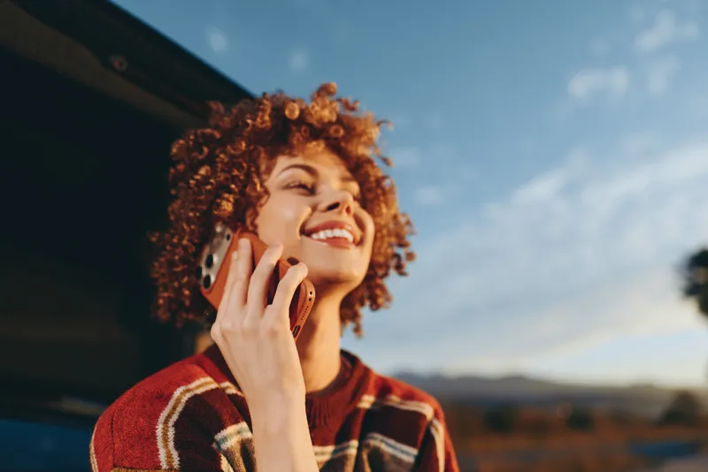 Smiling woman with curly hair outdoors, holding a phone receiver near her ear. Plaid sweater in soft daylight. Lifestyle moment for communication, joy, and self expression today.