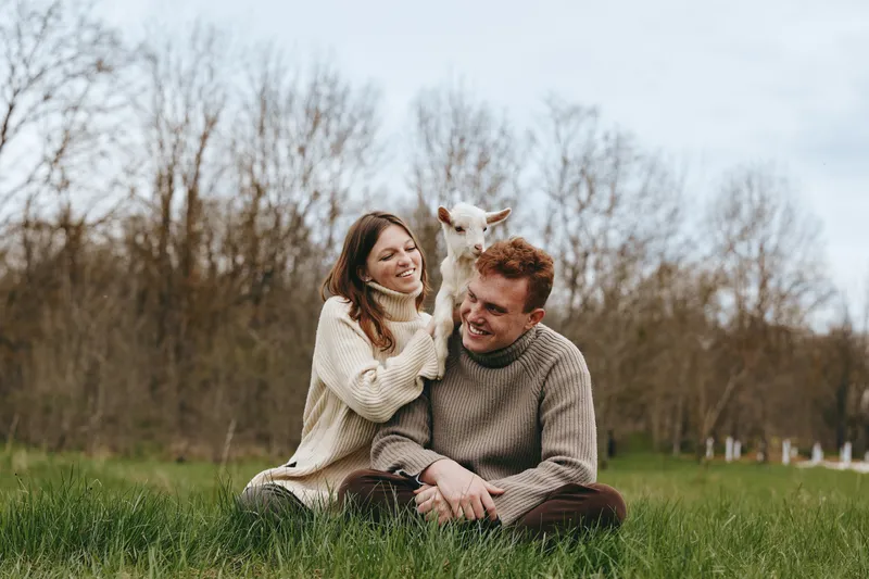 A cheerful couple sits on lush grass in an open meadow, a playful goat kid nearby. A candid lifestyle moment capturing companionship, rural charm, and gentle animal joy.