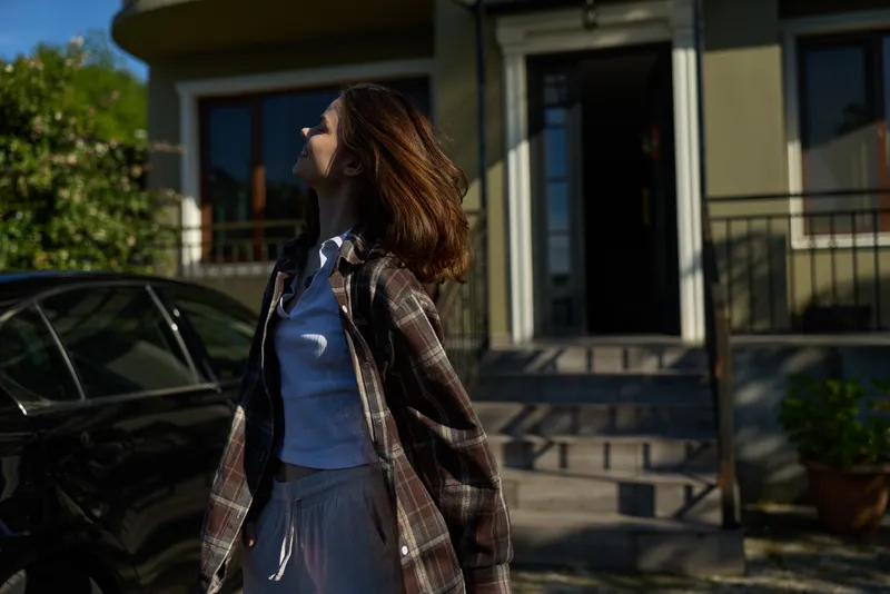 A woman in casual fashion stands outdoors in bright daylight on a residential porch, smiling with confidence, showcasing a lifestyle moment of everyday warmth and relaxed mood