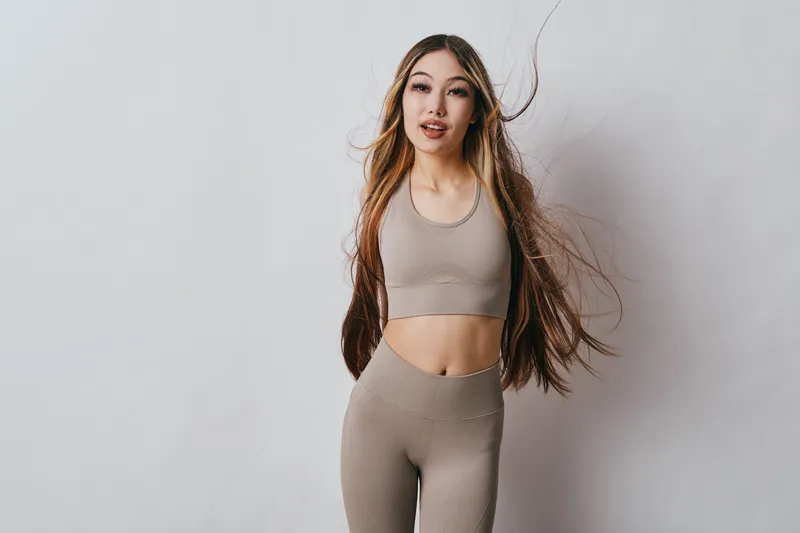 Young woman with long hair wearing beige sportswear posing on white background for fitness, health, wellness, and active lifestyle concept in studio lighting.