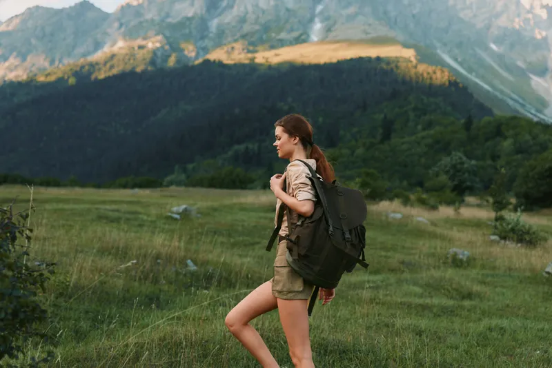 Cheerful woman hiking in a meadow with a large backpack, mountains in the distance, sunlight on grass, relaxed stride, outdoor lifestyle moment, natural colors and textures