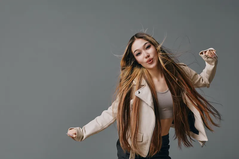 Woman with long hair wearing beige jacket and crop top posing on gray background, motion captured with playful expression and casual style in studio setting.