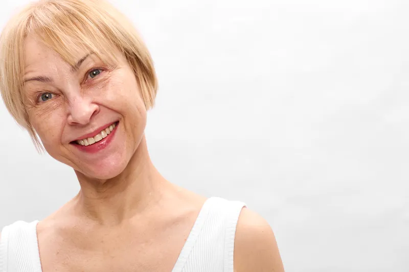 woman senior smiling portrait blonde studio closeup white background, relaxed expression, soft light, gentle lines, concept of aging well, positive wellbeing