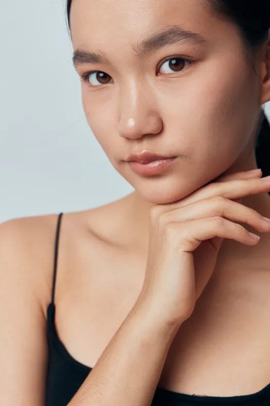Young woman with minimal makeup and natural skin posing in closeup portrait, hand gently touching chin. Calm expression with smooth complexion and focused eyes on light background.