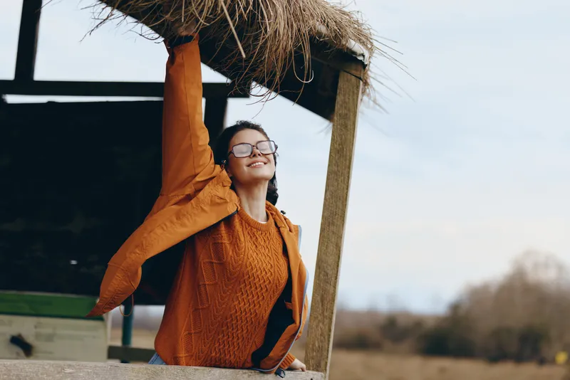 Woman lifestyle outdoors, smiling behind straw shelter, orange sweater and glasses, arms raised in relaxed pose, natural daylight, breezy air, countryside background