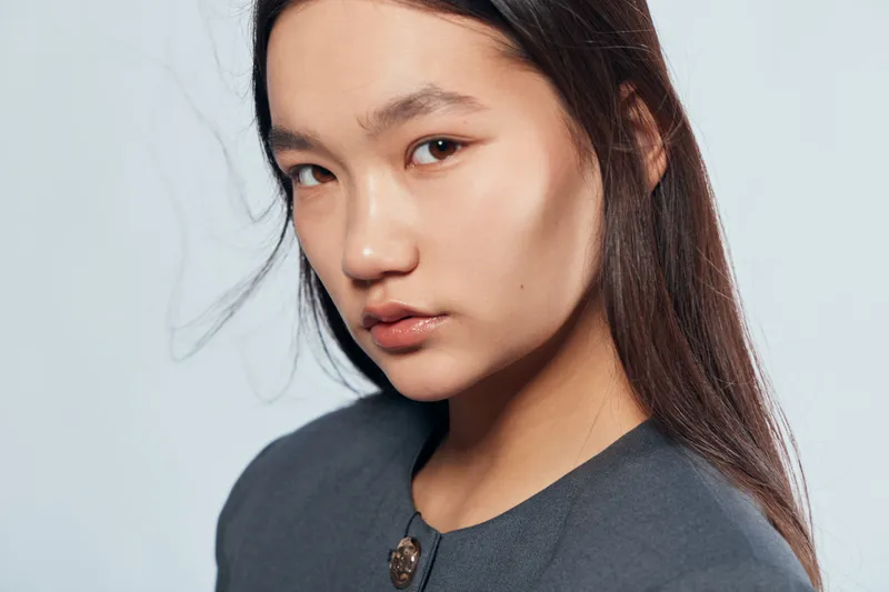 Young woman with long hair and natural makeup posing in studio with neutral expression. Female model wearing dark clothing with soft lighting on light background.
