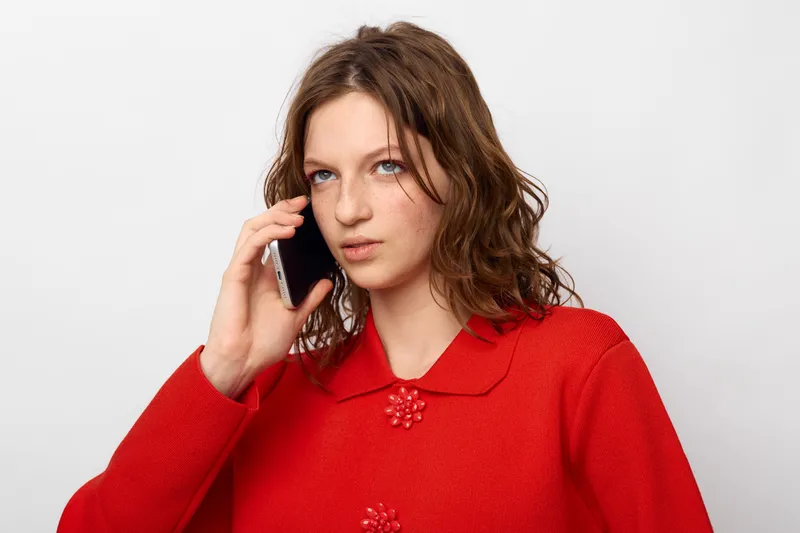 Young woman with curly hair wearing red sweater talks on smartphone against white background. Concept of communication, technology, casual style, and modern lifestyle.