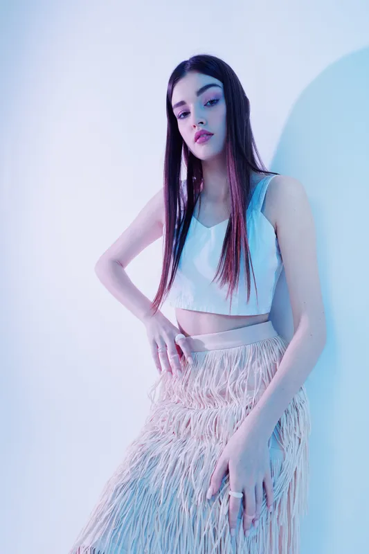 Young woman with long hair poses confidently in a white crop top and textured skirt under cool studio lighting. Concept of woman beauty fashion with modern style and elegance.