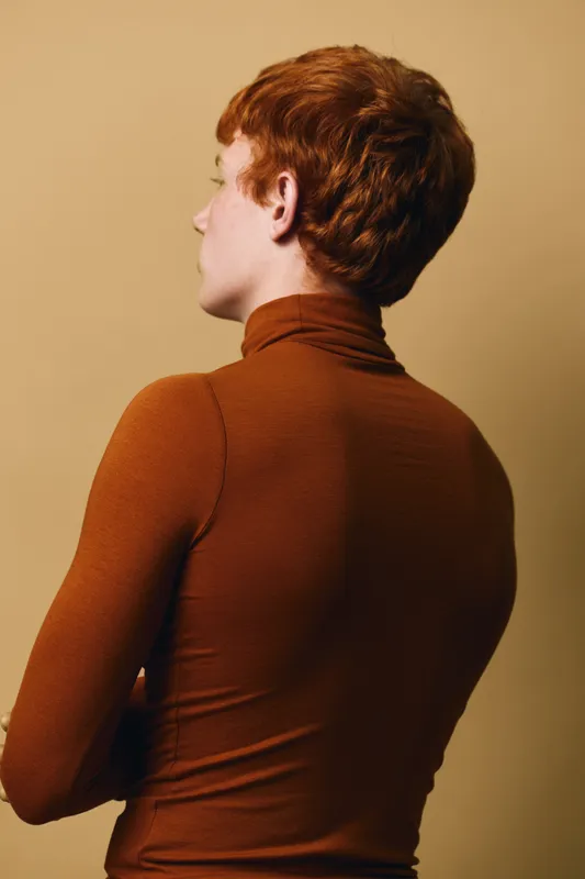 Profile view of a person with short red hair wearing a brown turtleneck top against a beige background. The subject is facing left with folded arms, showing a calm stance.