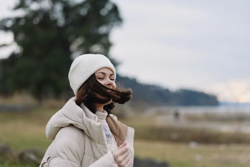 Winter scene with a woman in an outdoor coat and beanie, hair blown by wind, standing in a vast outdoor landscape under cloudy skies and calm atmosphere.