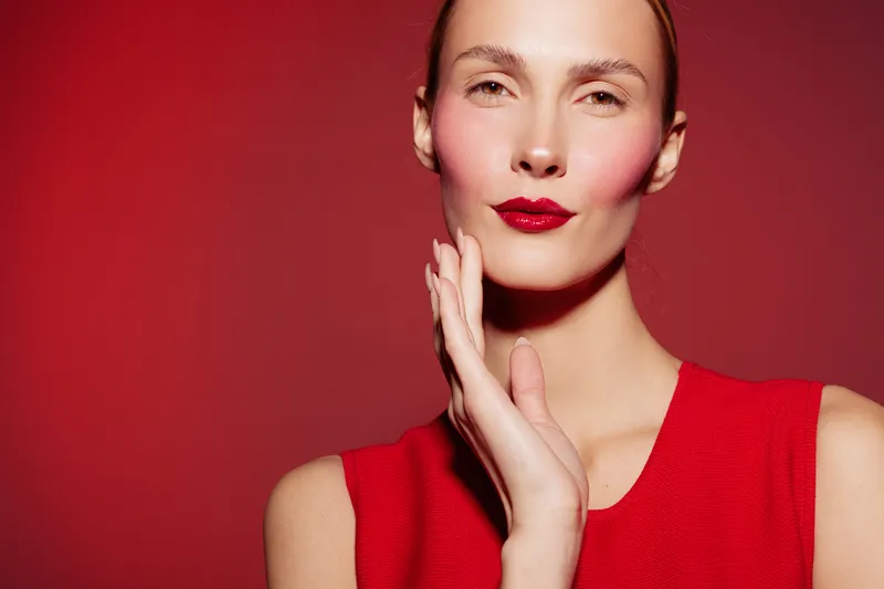 Close-up portrait of a confident woman with red lipstick and blush wearing a red sleeveless top against a red background. Concept of beauty, fashion, makeup, and elegance.