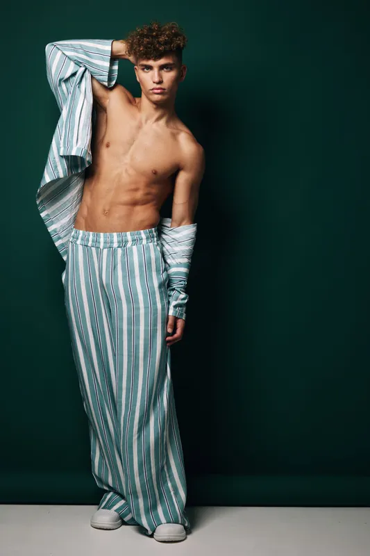 Young man with curly hair posing shirtless wearing striped loose pants and matching open shirt against dark background in studio fashion portrait.