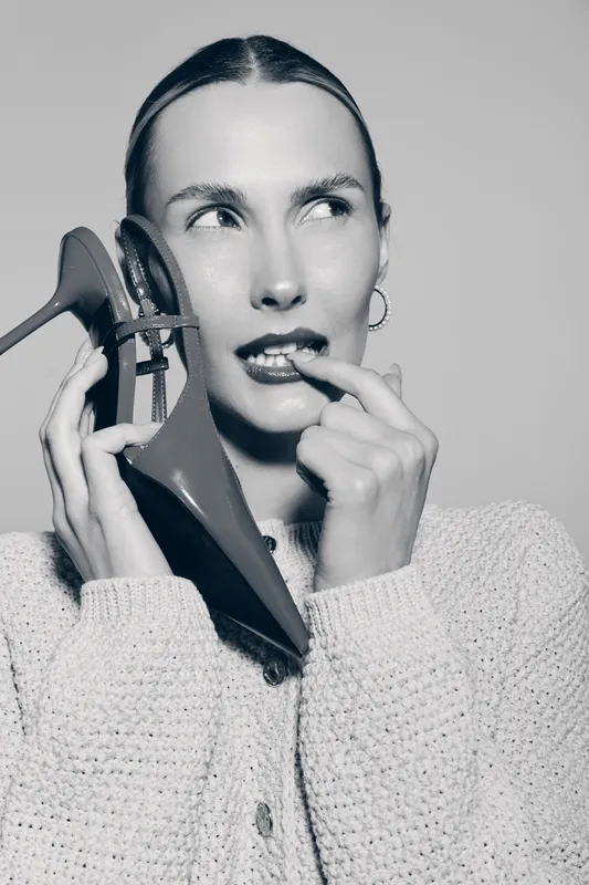 Young Caucasian woman with fair skin holds high heel shoe near face and bites finger in thoughtful pose. Concept of fashion, style, and beauty with monochrome background.
