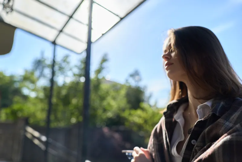 A thoughtful woman in casual plaid shirt stands outdoors under a glass canopy, sunlight on her face, looking away. A lifestyle moment focused on calm, reflection, and everyday serenity