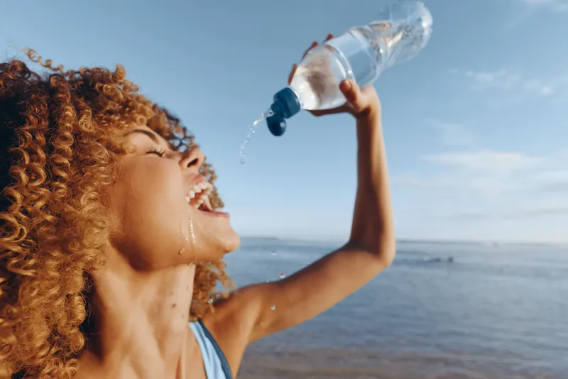 Curly haired woman drinks water from a plastic bottle near the ocean, smiling in bright summer daylight, showing hydration and wellness, outdoor refreshment by the sea, after travel