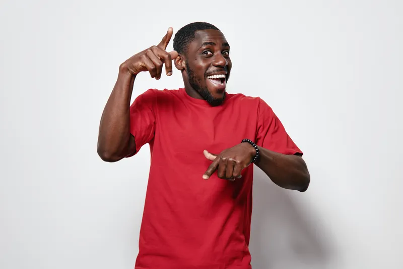 African man with dark skin wearing red t shirt smiling and pointing fingers up and down on white background. Happy black male showing directions with hands and joyful expression.