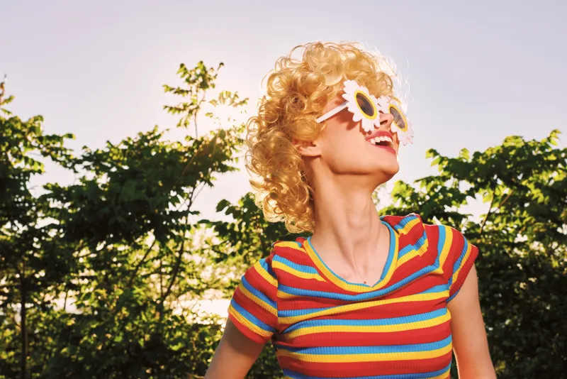 Cheerful woman outdoors, wearing a colorful striped top and quirky sunglasses, sunlit moment, carefree pose, vibrant colors, natural greenery background in a lively summer setting