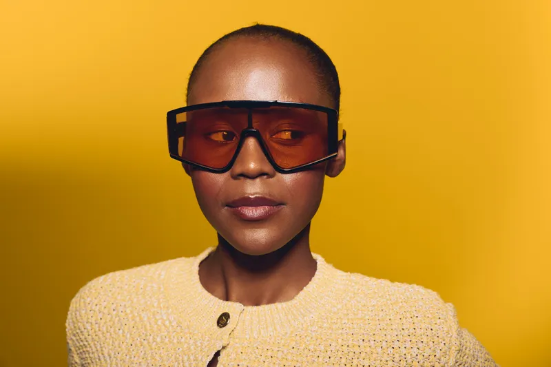 Portrait of confident dark skinned African woman wearing oversized sunglasses and yellow sweater against bright yellow background. Concept of fashion, style, and modern beauty.
