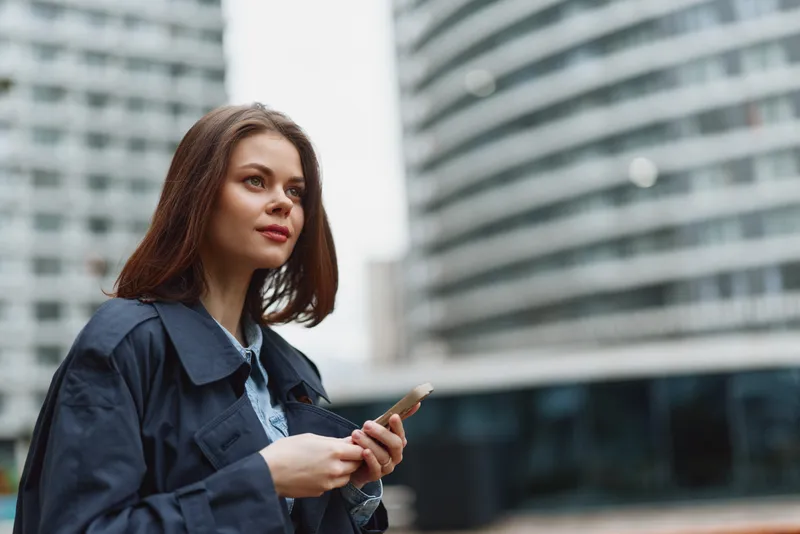 Young woman with long hair using smartphone outdoors in urban environment. She wears a dark jacket and looks thoughtfully into the distance. Modern city background.