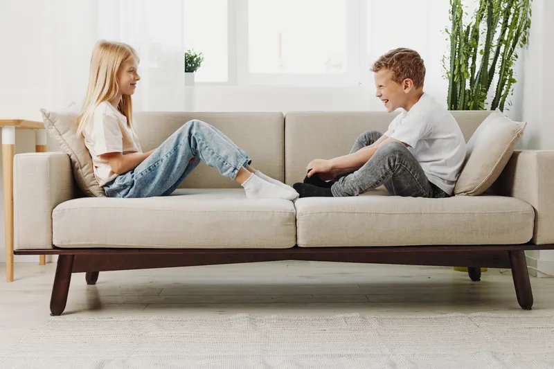 Happy young boy and girl sitting opposite each other on beige sofa in bright modern living room, smiling and enjoying playful conversation. Concept of friendship, childhood, and relaxed home