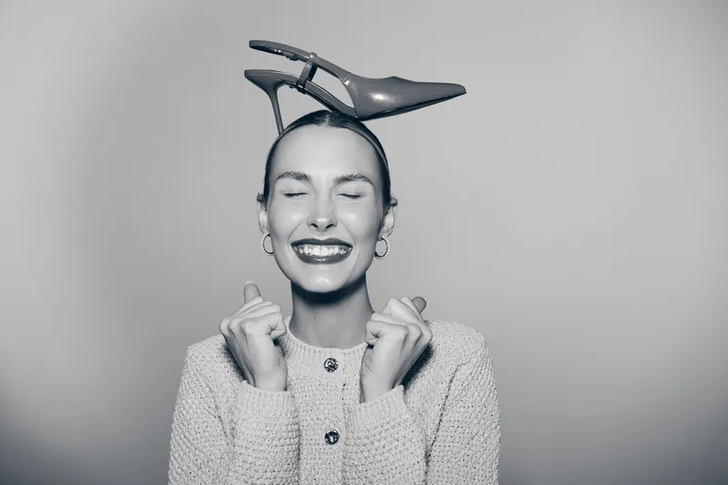 Happy Caucasian woman with light skin smiles with closed eyes and holds a high heel shoe on her head in studio. Concept of joy, fashion, and playful mood.