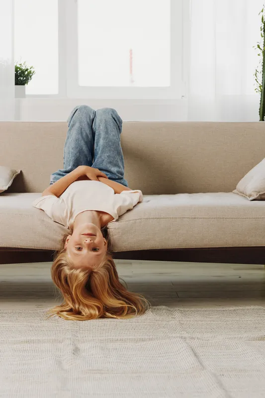 Blonde child lying upside down on beige sofa in bright living room with large windows. Casual denim jeans and white shirt create relaxed mood. Concept for childhood, playfulness, and home comfort.