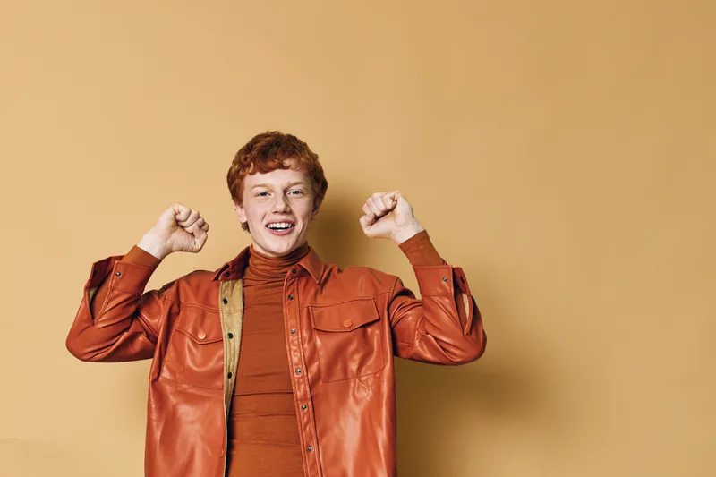 Young man with red hair wearing orange jacket and shirt is smiling with raised fists on beige background for celebration and happiness.