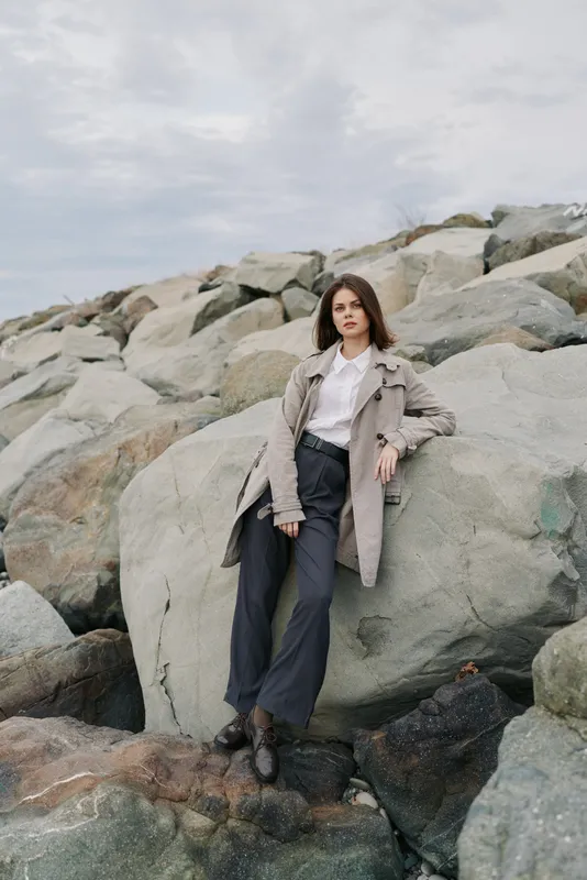 Young woman in stylish coat and wide pants poses confidently on large rocks under cloudy sky. Fashion photo shoot concept with natural background and casual modern outfit.