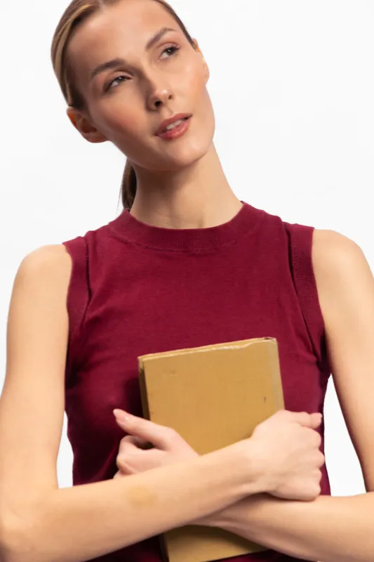 Thoughtful young woman in red sleeveless top holding a book with both hands, looking away on white background. Concept of education, contemplation, and knowledge.