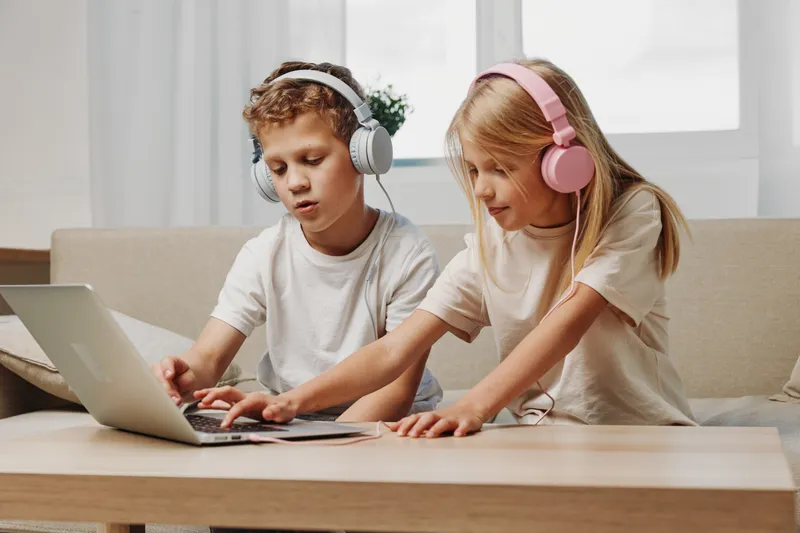 Two children with headphones use laptop together at home, focused on learning and digital interaction on sofa in bright living room.