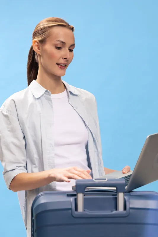 Young woman with casual outfit standing with suitcase and laptop against blue background. Concept of travel planning, technology use, and vacation preparation.