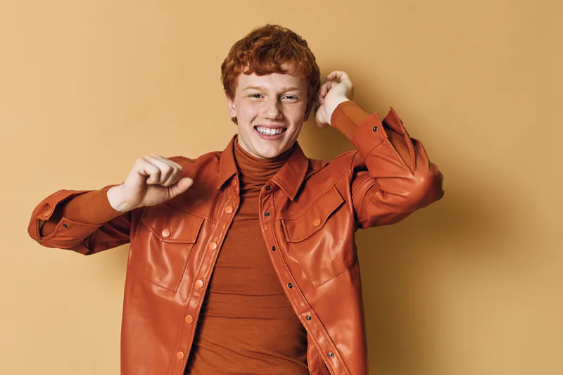 Young man with red hair wearing orange leather jacket and turtleneck smiling and dancing against beige background indoors with happy expression and casual style.