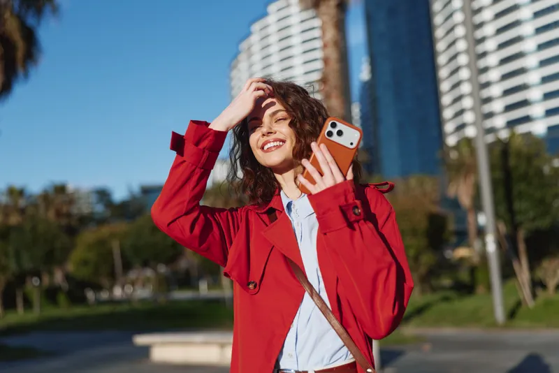 A young woman in a red coat holds a smartphone and smiles while standing outdoors in a sunny urban park with modern buildings in the background, enjoying a bright day.