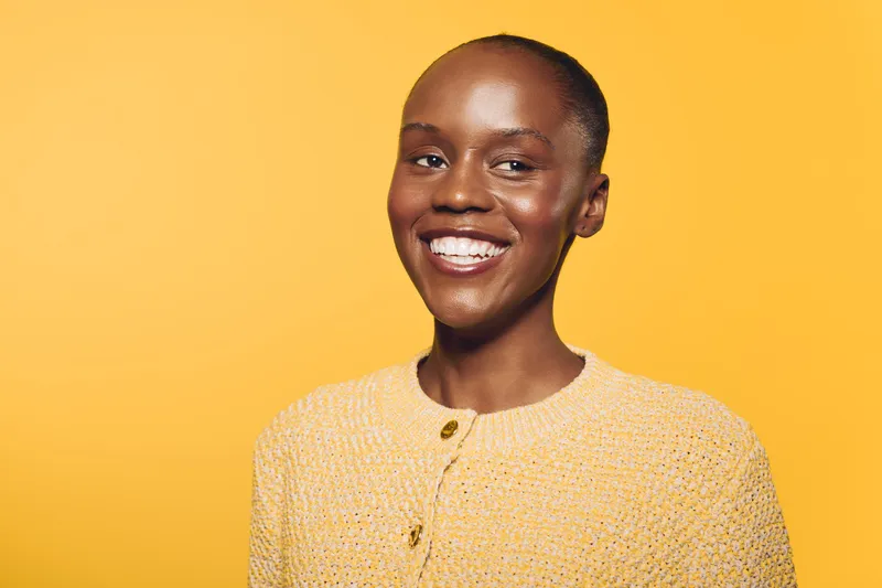 Smiling black woman with dark skin wearing a yellow sweater poses against a yellow background. The image conveys happiness, confidence, and natural beauty in a bright studio setting.
