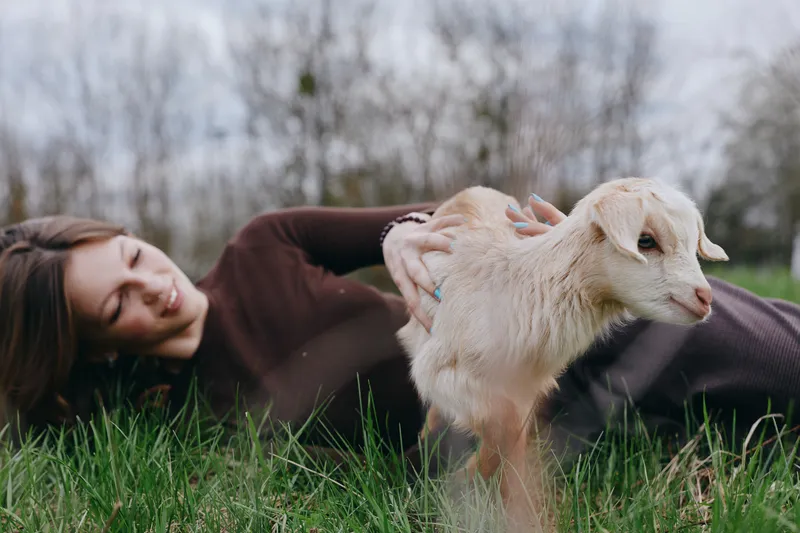 Woman lying on grass with a playful goat kid nearby, an outdoors lifestyle shot that captures friendship between human and animal, natural light, calm nature, and a simple countryside atmosphere.